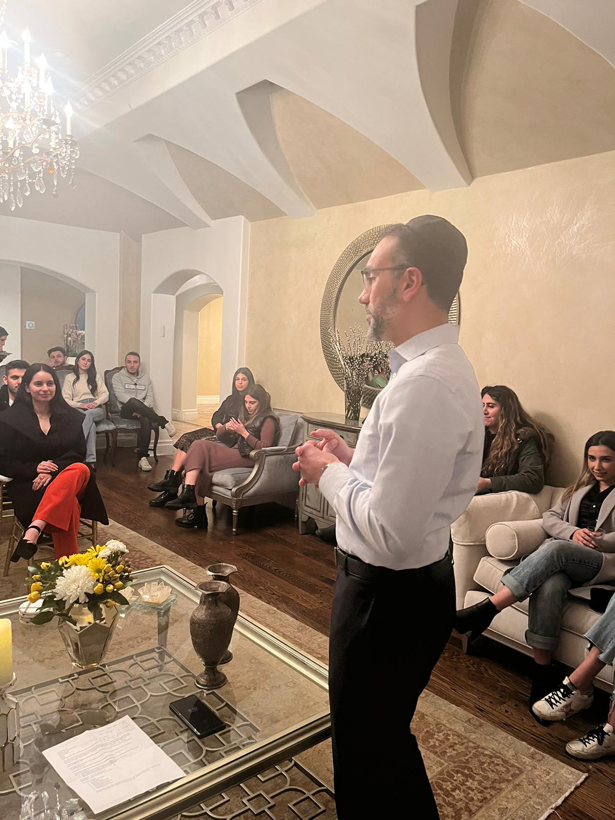 Rabbi Nissan mid-gesture, teaching a small seated group in a warm living room